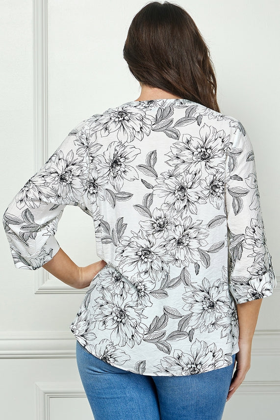 A woman with long brown hair, seen from behind, wears the Plus Floral Button Blouse featuring black floral prints and relaxed fit, paired with blue jeans. She stands in front of a light-colored wall with molding details.