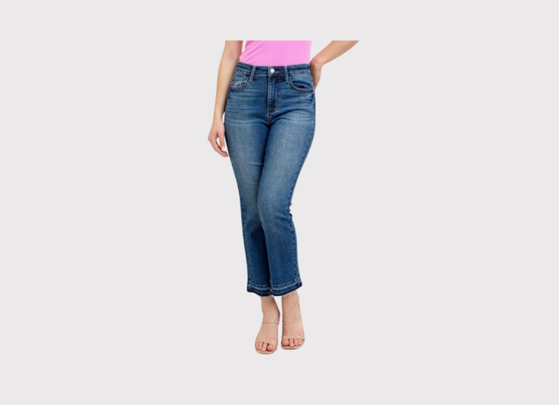 A person wearing a bright pink t-shirt and Judy Blue cropped jeans with a raw, frayed hem. The person is standing on a light background and also wearing light-colored open-toed sandals.