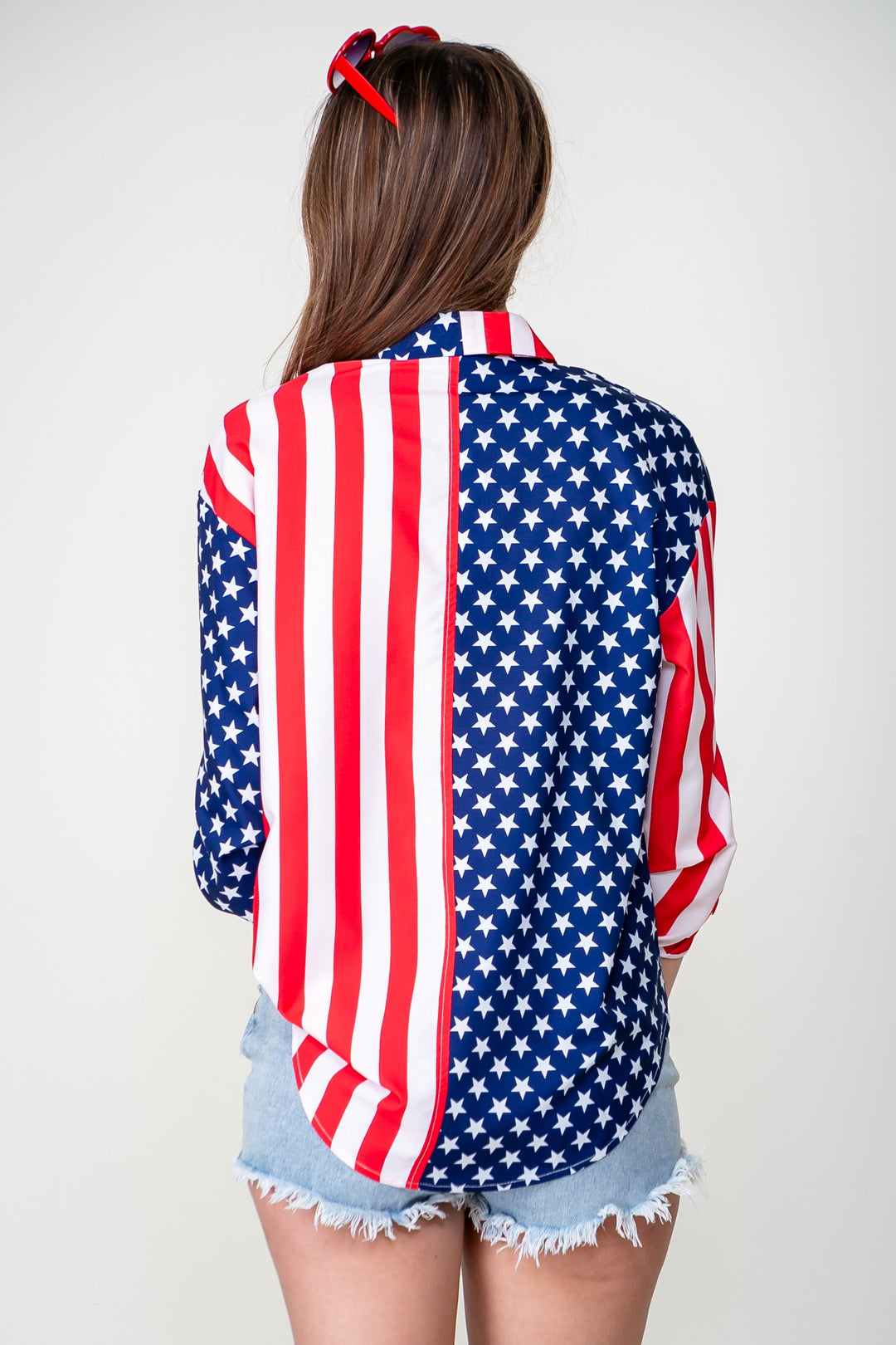 A person with long brown hair, wearing a Patriotic Button Shirt and light denim shorts, stands facing away. Their look is completed with a red bow headband for a classic touch.