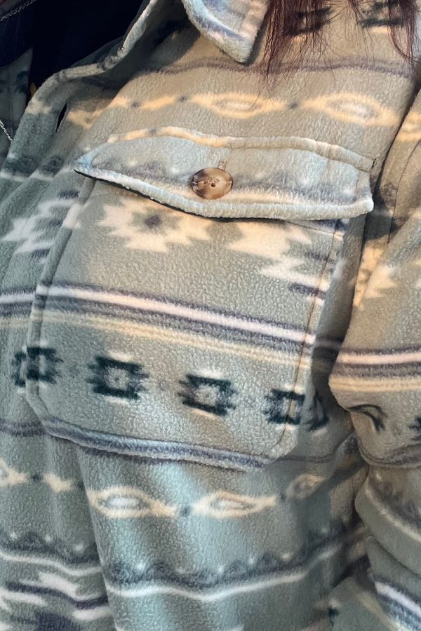 A close-up of the Southwestern Fleece Shacket highlights its buttoned chest pocket and soft fleece fabric, showcasing a geometric zigzag pattern in muted shades of green, beige, and gray.