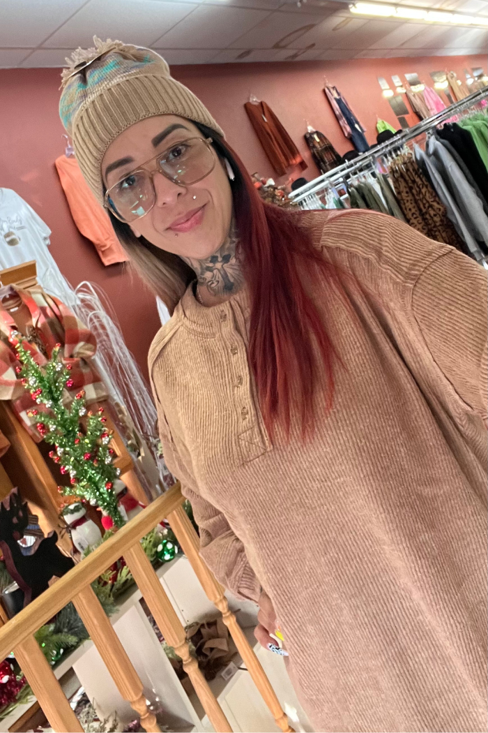 A woman with red-tinted hair, glasses, and a knit hat smiles in a holiday-decorated clothing store, wearing a tan Brushed Ribbed Henley Top with racks of clothes and a small Christmas tree in the background.