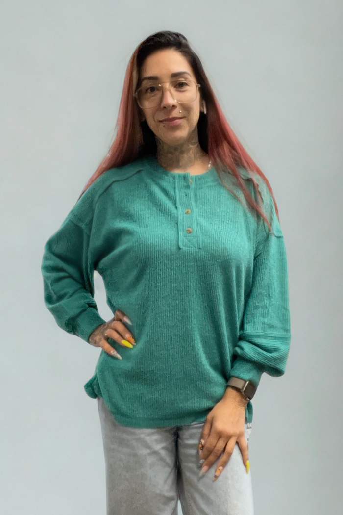 A person with long reddish-brown hair, glasses, and tattoos poses with one hand on their hip in a green Brushed Ribbed Henley Top and light-colored pants, standing against a plain light background for a relaxed look.