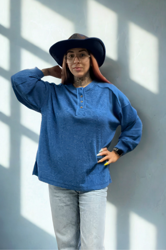 Wearing a blue Brushed Ribbed Henley Top, light jeans, and a wide-brimmed hat, a person stands against a light wall with window shadows, smiling and raising a hand to their hat in relaxed, cozy loungewear style.