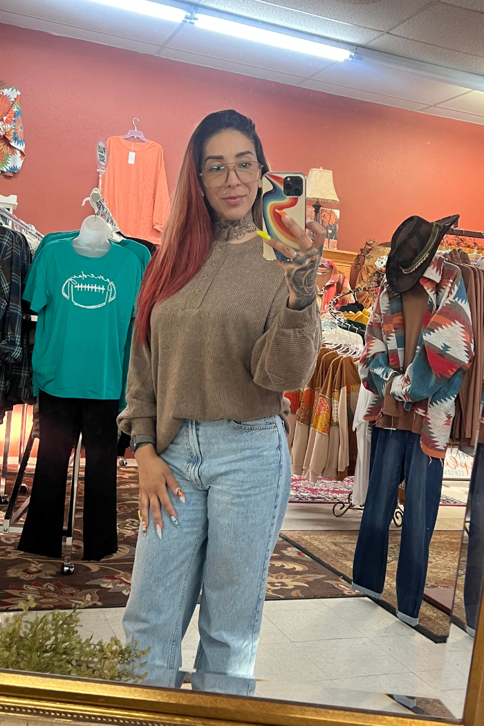 A woman with long, reddish hair and glasses takes a mirror selfie in a clothing store, wearing the Brushed Ribbed Henley Top in tan with light blue jeans. Colorful clothes and mannequins are visible in the background.