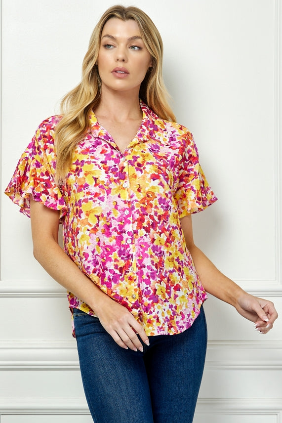 A woman with long, wavy blonde hair poses against a white wall in the Floral Button Blouse, a lightweight woven short-sleeve top featuring a collared neckline and vibrant pink, yellow, and white abstract floral pattern.