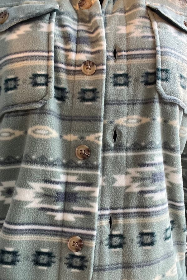 Close-up of the Southwestern Fleece Shacket, showcasing a blue, white, and gray geometric pattern. One button is missing, leaving an empty buttonhole in the center. The fabric appears soft and fleece-like.