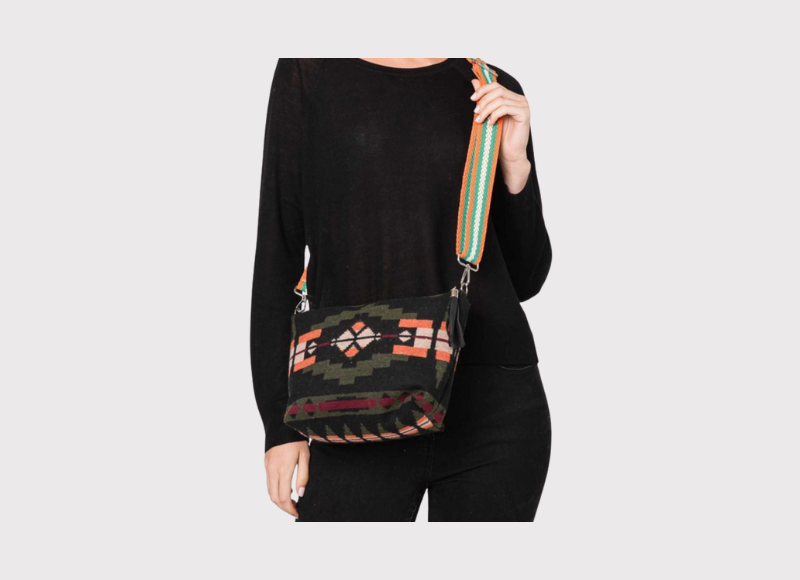A woman wearing a black long-sleeved top and black pants carries a crossbody bag with a dark green and orange Aztec pattern and a colorful striped shoulder strap featuring shades of orange, green, blue, and white.