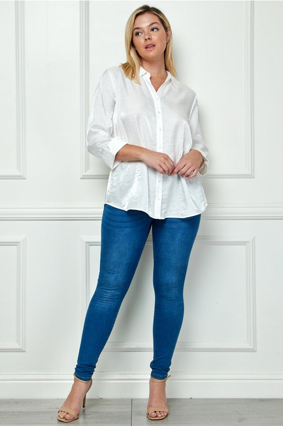 A woman with blonde hair poses confidently against a white paneled wall, wearing the Plus Shimmer Collar Blouse in white with an oversized fit, paired with blue skinny jeans and beige heels.