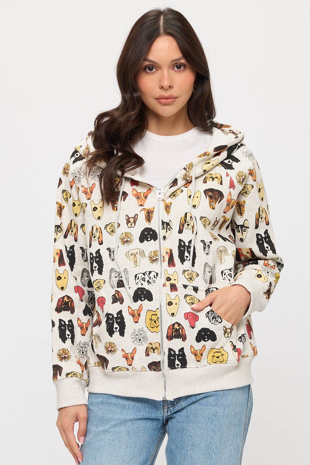 A woman with long, wavy dark hair models the COLORFUL DOG FACES PRINT ZIP UP HOODIE against a white background, pairing the playful dog lover apparel with light blue jeans.