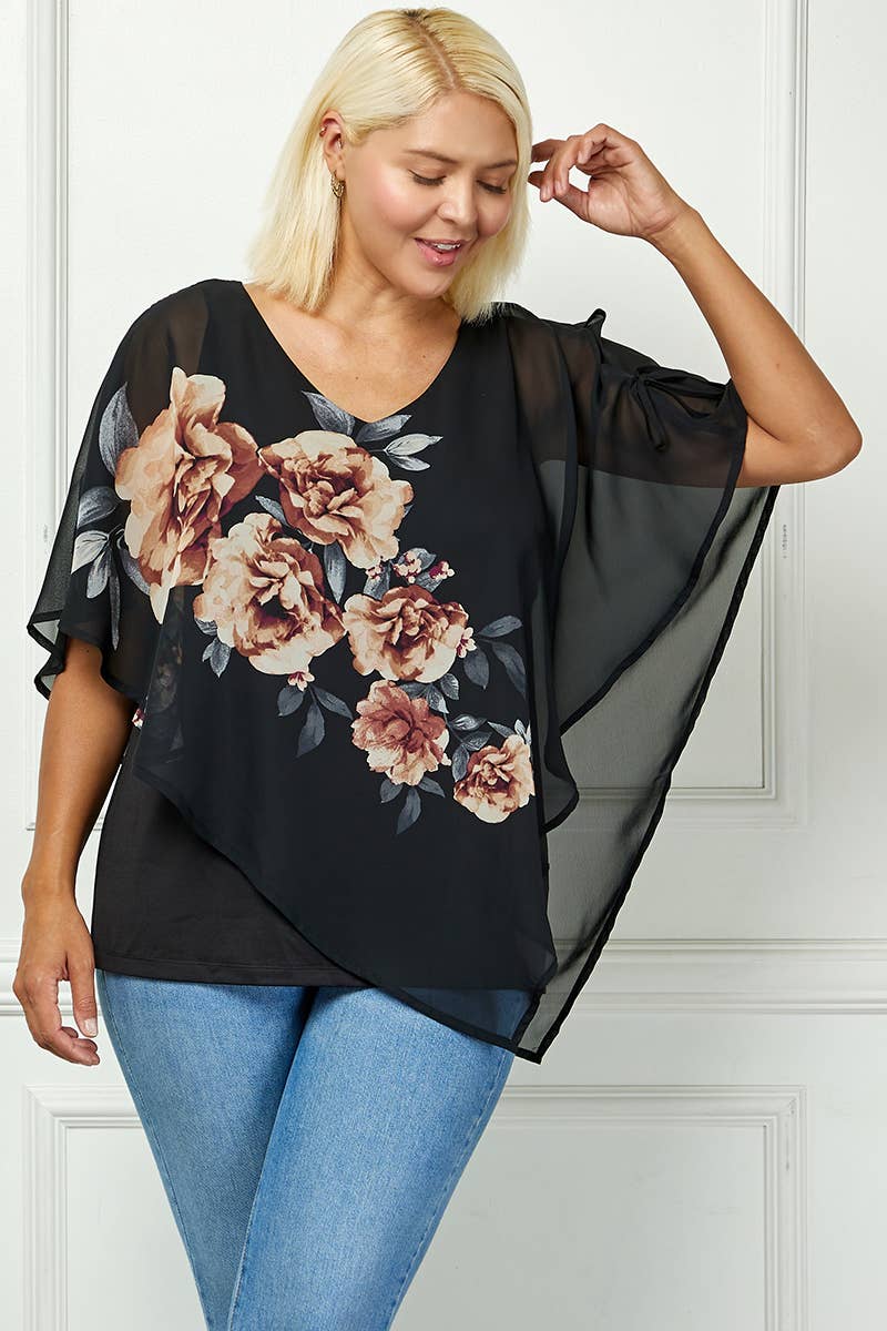 A woman with light blonde hair wears the Plus Blooming Poncho Blouse over a black tank and blue jeans, smiling and touching her hair as she stands before a white paneled wall.