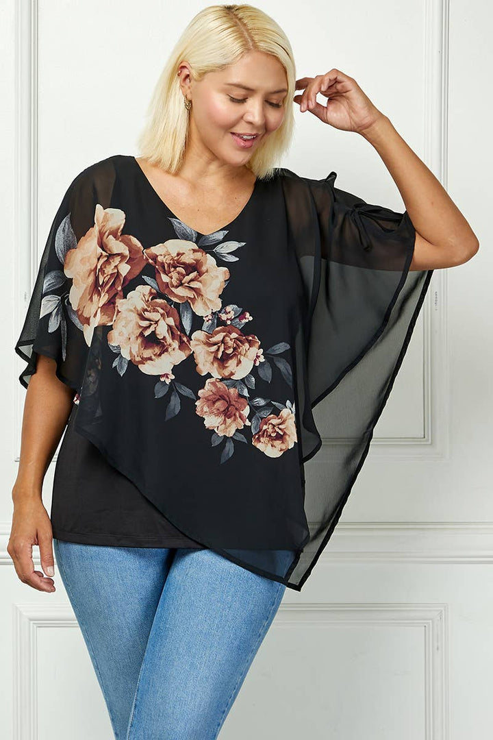 A woman with light blonde hair wears the Plus Blooming Poncho Blouse over a black tank and blue jeans, smiling and touching her hair as she stands before a white paneled wall.