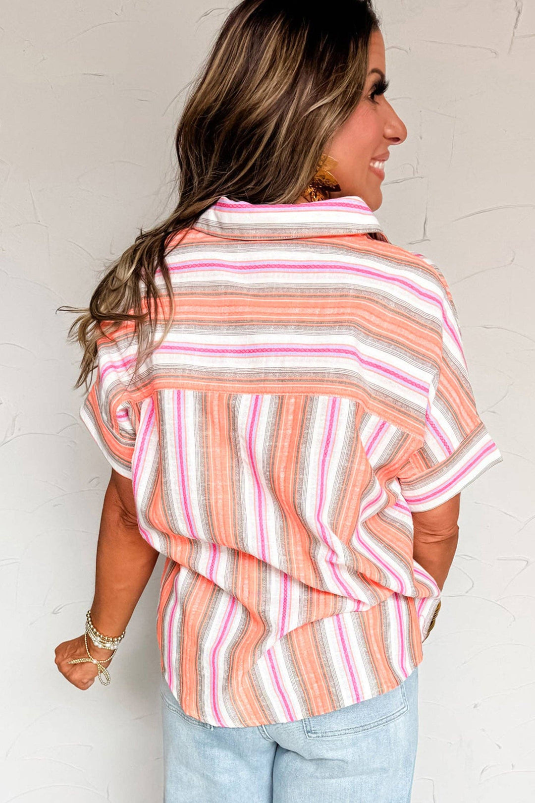 A woman with long, wavy brown hair smiles with her back to the camera, wearing the Multicolor Stripe Shirt in orange, pink, and white. The short sleeve button-up pairs with light blue jeans for a comfortable, casual look.