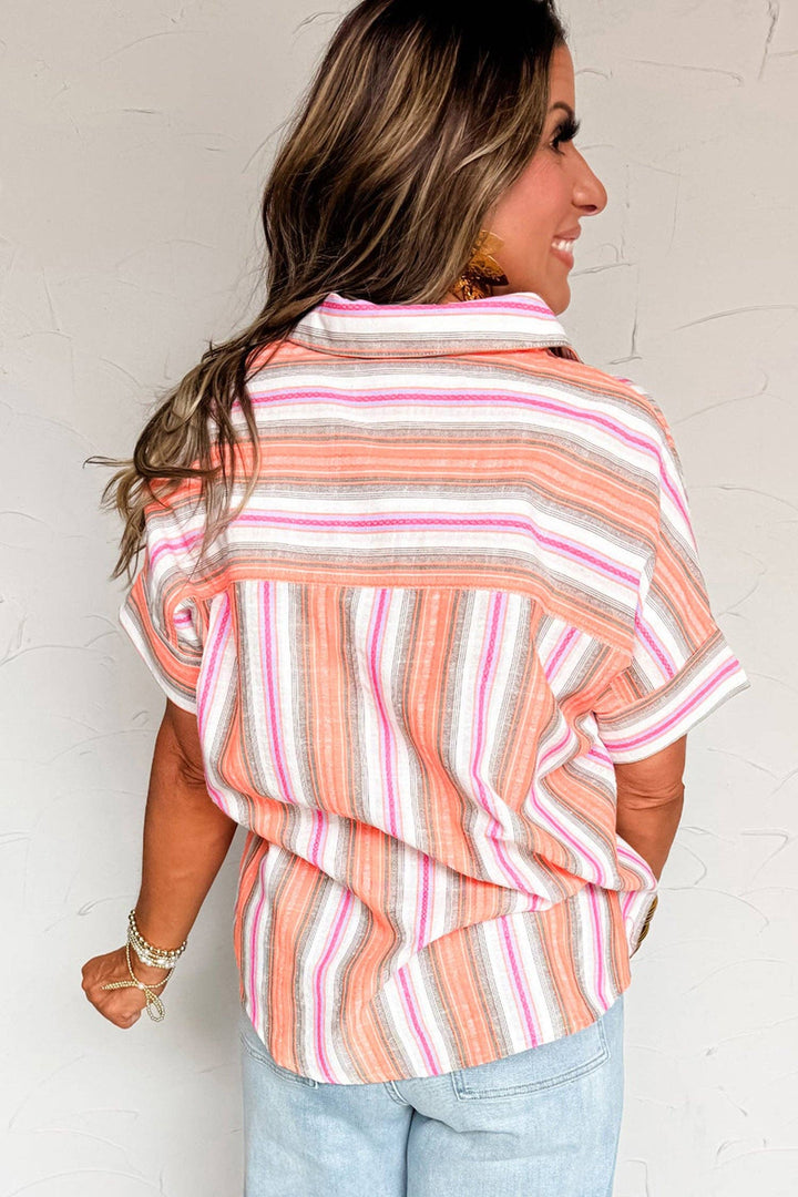 A woman with long, wavy brown hair smiles with her back to the camera, wearing the Multicolor Stripe Shirt in orange, pink, and white. The short sleeve button-up pairs with light blue jeans for a comfortable, casual look.