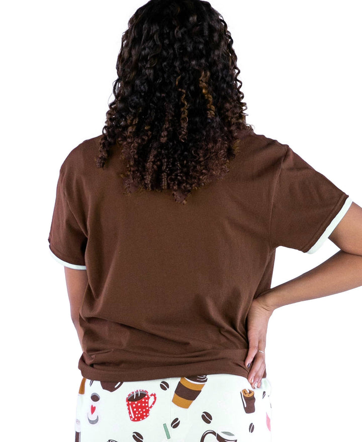 A person with curly hair, seen from behind, wears a Dog Mom, Cat Nap & More Pajama Top in brown cotton and patterned pajama pants with novelty coffee cup and donut prints. Their left hand rests on their hip.