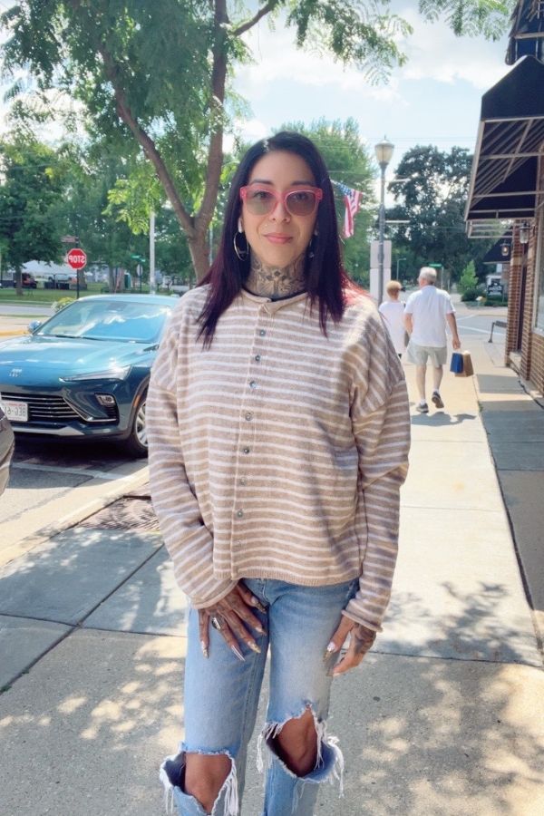 A woman with long dark hair, pink glasses, and tattoos is standing on a sunny sidewalk wearing the Striped Button Front Sweater and ripped jeans, with trees and parked cars in the background.