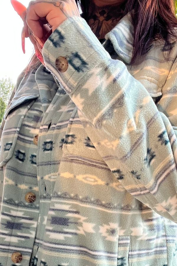 A person wearing the Southwestern Fleece Shacket in a light blue geometric pattern with large brown buttons, holding the collar. Part of their hand and long fingernail are visible.