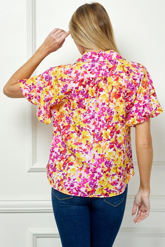 A woman with blonde hair, wearing the Floral Button Blouse featuring pink, yellow, and orange florals with a collared neckline and blue jeans, stands touching her temple in front of a white paneled wall.