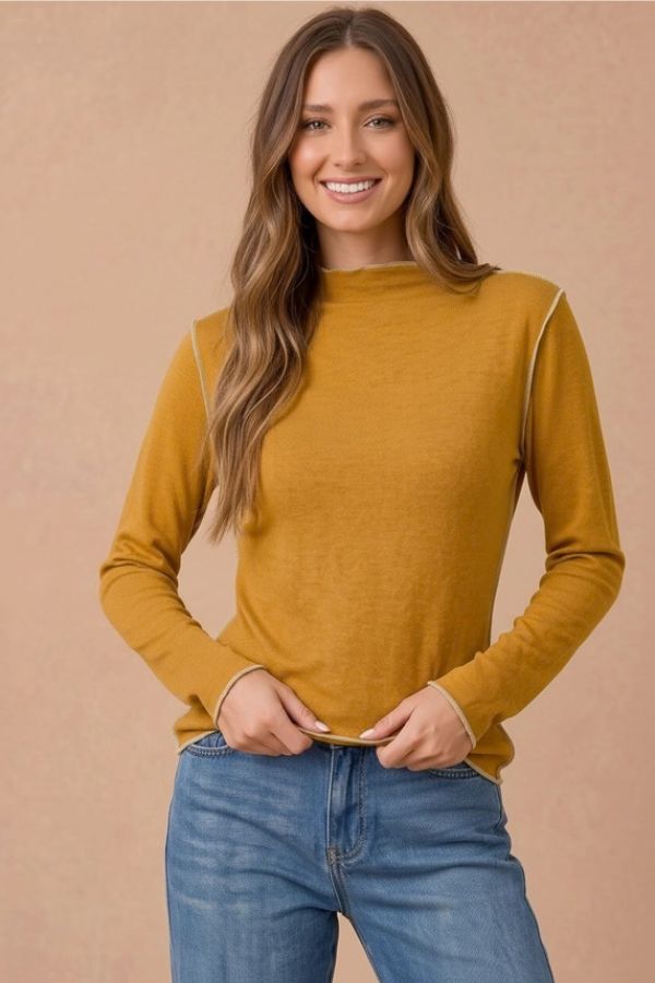 A woman wears a mustard yellow Frilled Mock Neck Hacci top with exposed light-colored seam detailing and blue jeans.