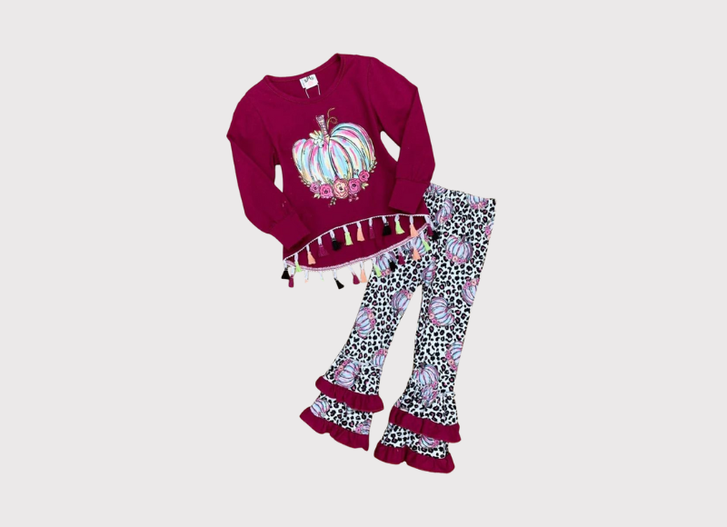 A children's two-piece fall outfit featuring a burgundy long-sleeved top with a colorful, decorated pumpkin graphic and multi-colored tassels along the hem, paired with ruffled bell-bottom pants in a black and white leopard print with burgundy ruffled cuffs.