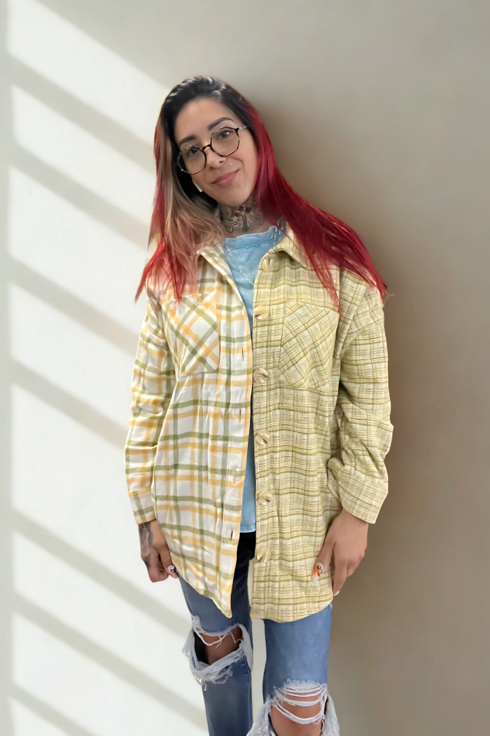 A person with red-tipped, dark hair and glasses smiles while wearing an unbuttoned yellow, white, and green plaid shacket over a light blue top, with ripped blue jeans. They have visible tattoos on their neck and arms. 