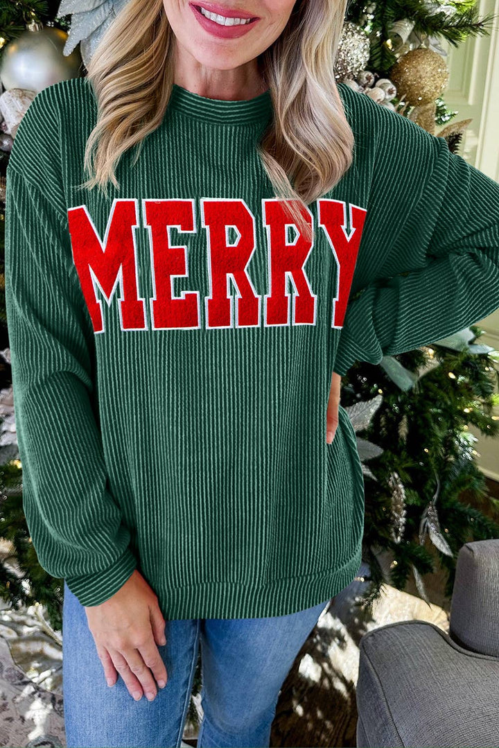 A woman stands by a Christmas tree, smiling, wearing the Merry Graphic Top—a green ribbed sweater featuring bold red "MERRY" lettering across the front. Only her upper body and part of her face are visible, showcasing this charming festive top that’s perfect for holiday gatherings.