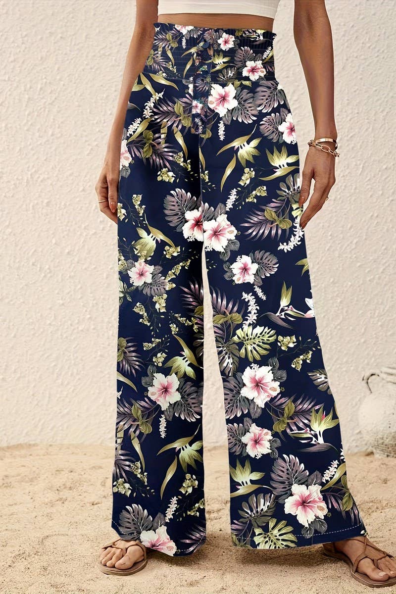 A person stands on sand wearing Tropical Wide Leg Pants featuring a floral print and high-rise elastic waistband, paired with sandals, a crop top, and bracelets, set against a textured light wall background.
