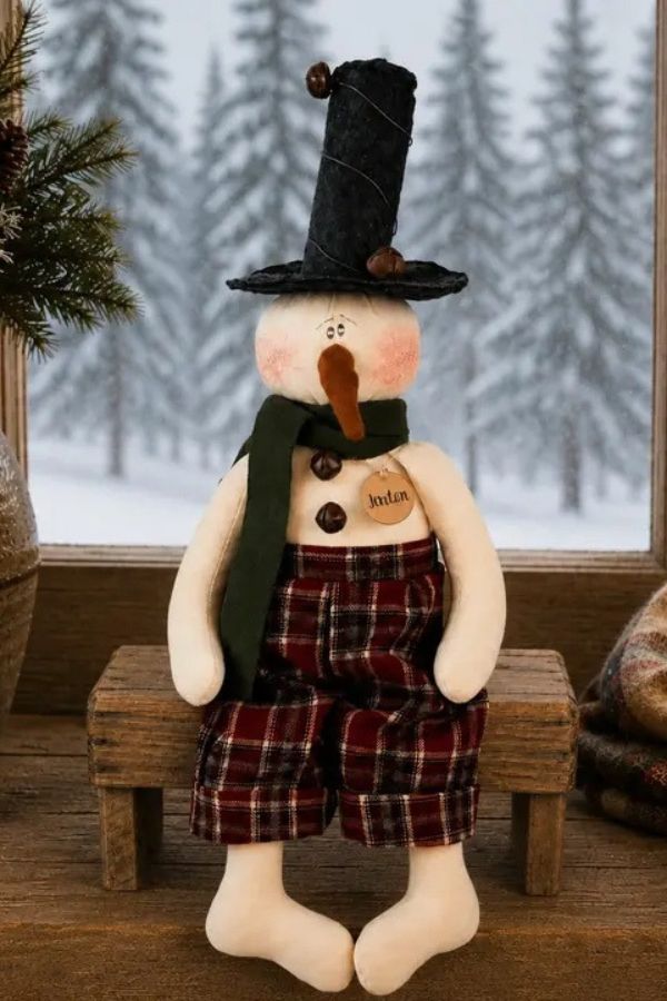 A Jaxton Jingle Bell Snowman Shelf Sitter wearing a black top hat, dark green scarf, and plaid pants, resting on a small wooden bench in front of a snowy window.