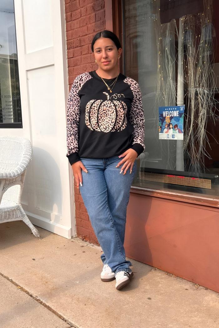 Woman wearing a black sweatshirt with a pumpkin design, standing on a sidewalk.