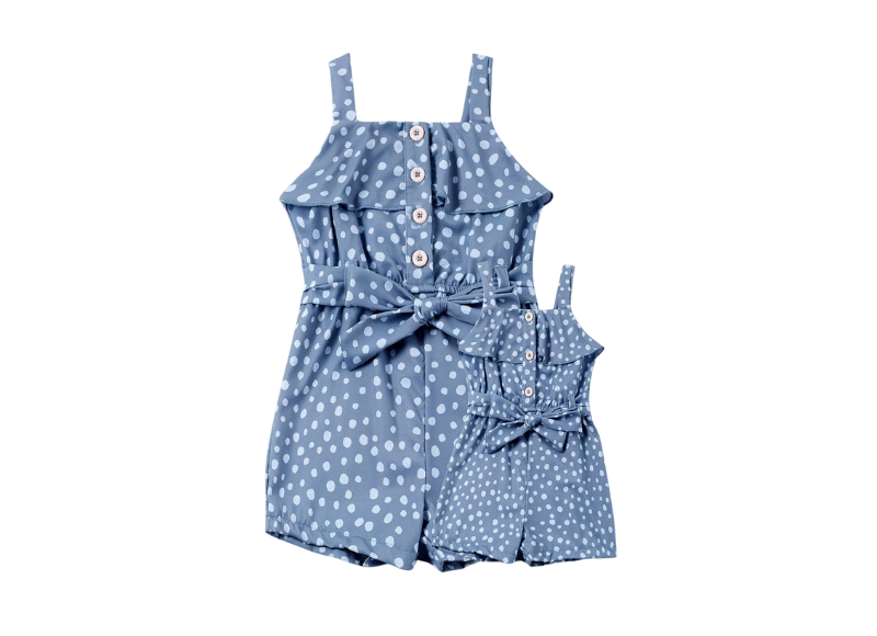 The image shows a matching set of jumpsuits, one for an adult and one for a child, both featuring a blue background with white polka dots. They have ruffled straps, button details, and a tie waist. The smaller jumpsuit is designed for a doll or young child, while the larger one is for an adult, creating a "Mommy & Me" style.