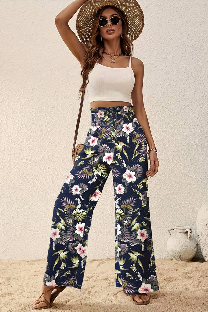 A woman stands on sand against a light textured wall, wearing sunglasses, a white crop top, a straw hat, and the Tropical Wide Leg Pants—high-rise with an elastic waistband and a tropical floral print on navy.