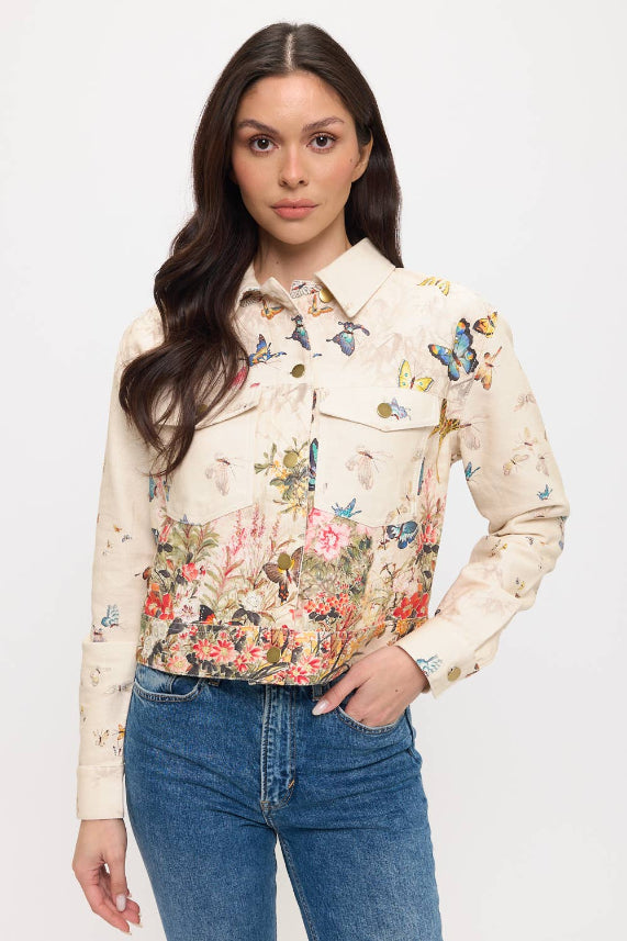 A woman with long brown hair models the Butterfly Floral Jacket, a cropped piece with colorful florals and butterflies, paired with blue jeans against a plain white background.