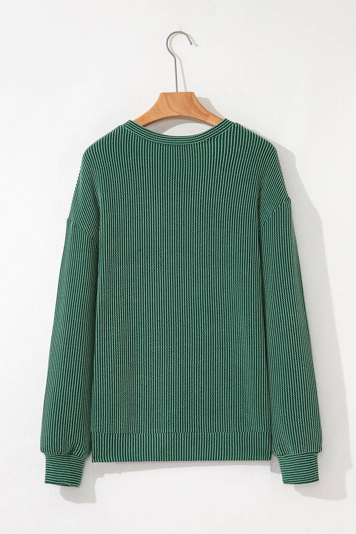 A green, long-sleeve Merry Graphic Top hangs on a wooden hanger against a white wall. Perfect for holiday gatherings or cozy winter wear, the ribbed-knit sweater is shown from the back.