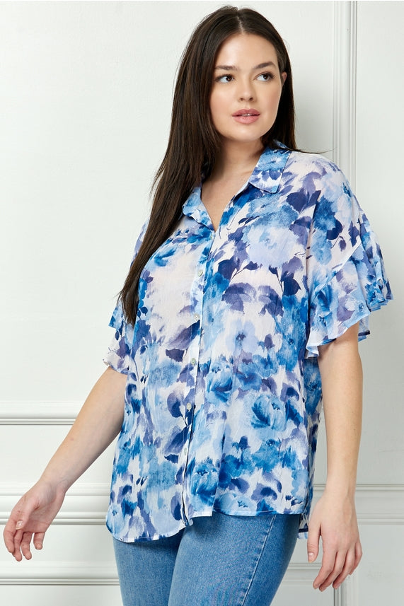 Woman with long dark hair wearing the Plus Floral Ruffle Blouse in a blue watercolor floral print and light blue jeans, standing against a white wall.
