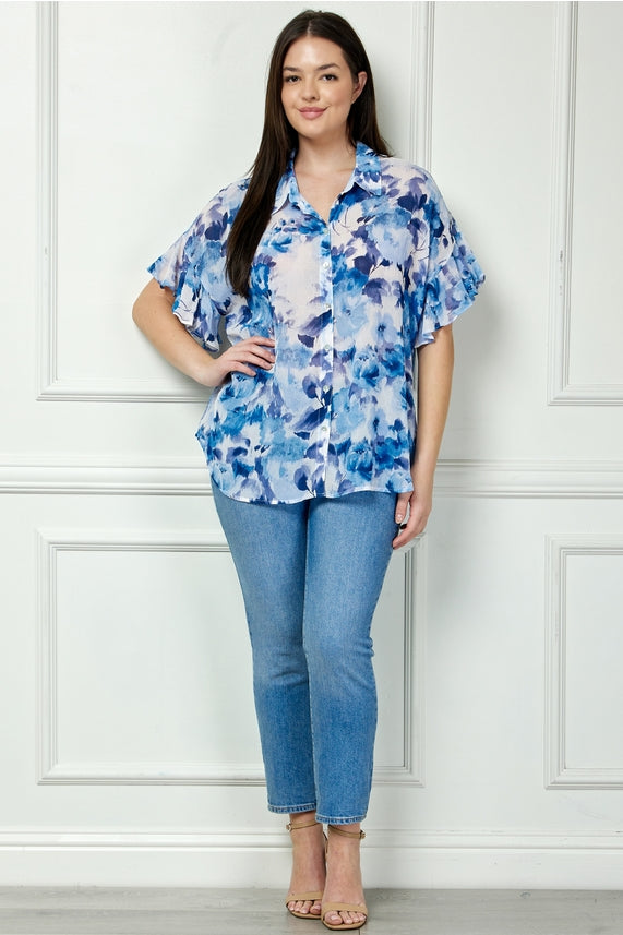 A woman with long dark hair stands indoors against a white wall, smiling slightly. She wears the Plus Floral Ruffle Blouse in a blue and white print, paired with light blue jeans and beige heeled sandals.