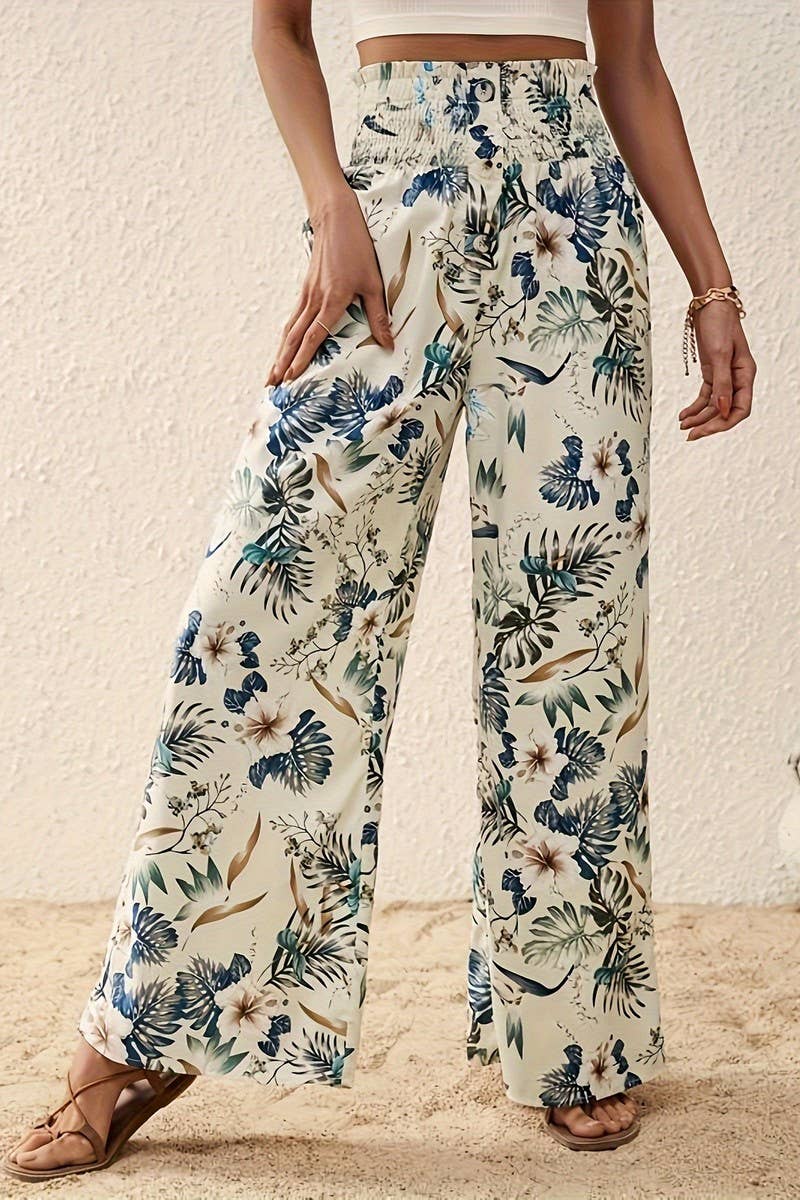 A woman, shown from torso to legs, stands on sand against a textured white wall wearing Tropical Wide Leg Pants featuring a green, blue, and beige floral print and a high-rise elastic waistband.