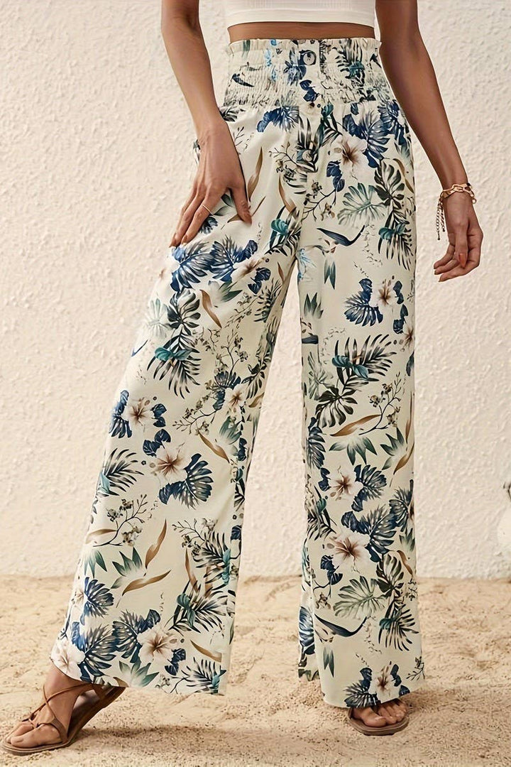 A woman, shown from torso to legs, stands on sand against a textured white wall wearing Tropical Wide Leg Pants featuring a green, blue, and beige floral print and a high-rise elastic waistband.