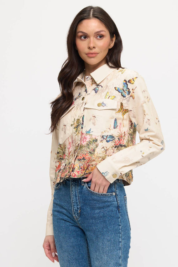 A woman with long dark hair wears the Butterfly Floral Jacket—featuring a cream color with a vibrant botanical print—paired with blue jeans, standing against a plain white background.