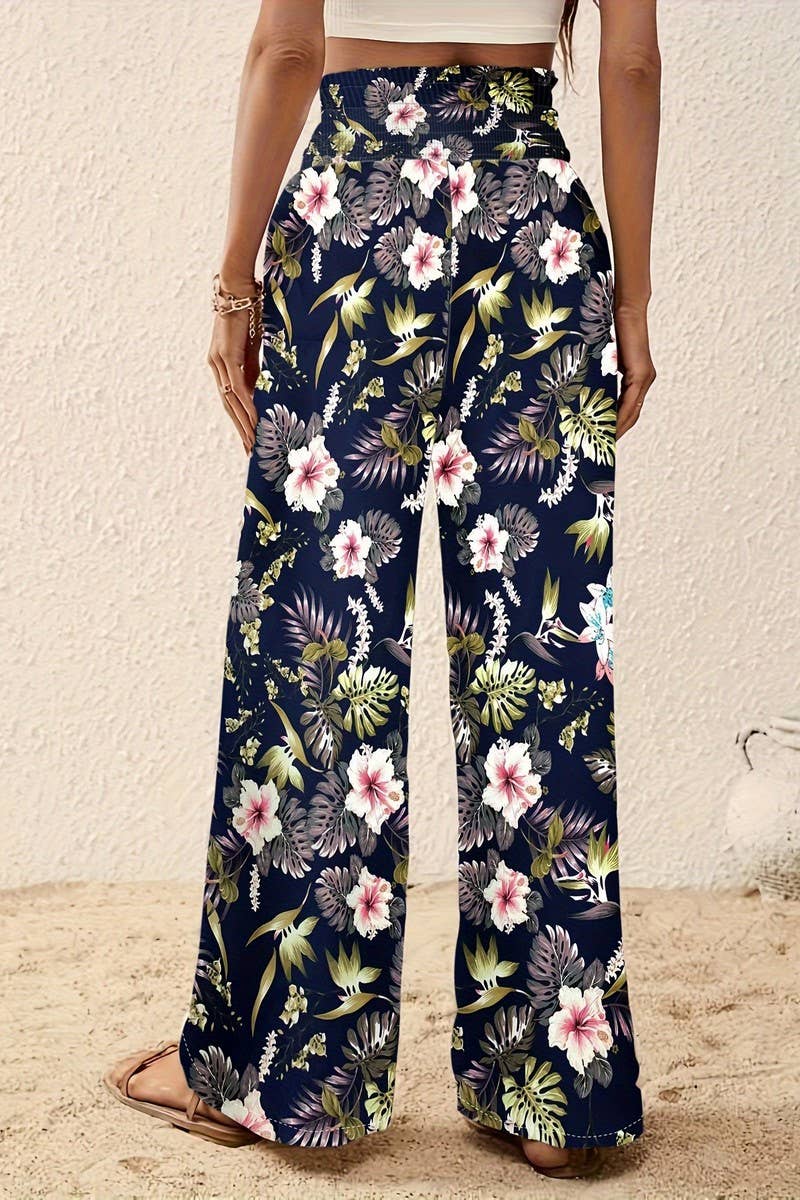 A woman stands on sand wearing the Tropical Wide Leg Pants, which feature a vibrant tropical floral print and a high-rise elastic waistband. Photographed from behind, she pairs them with a white top and sandals for a breezy look.