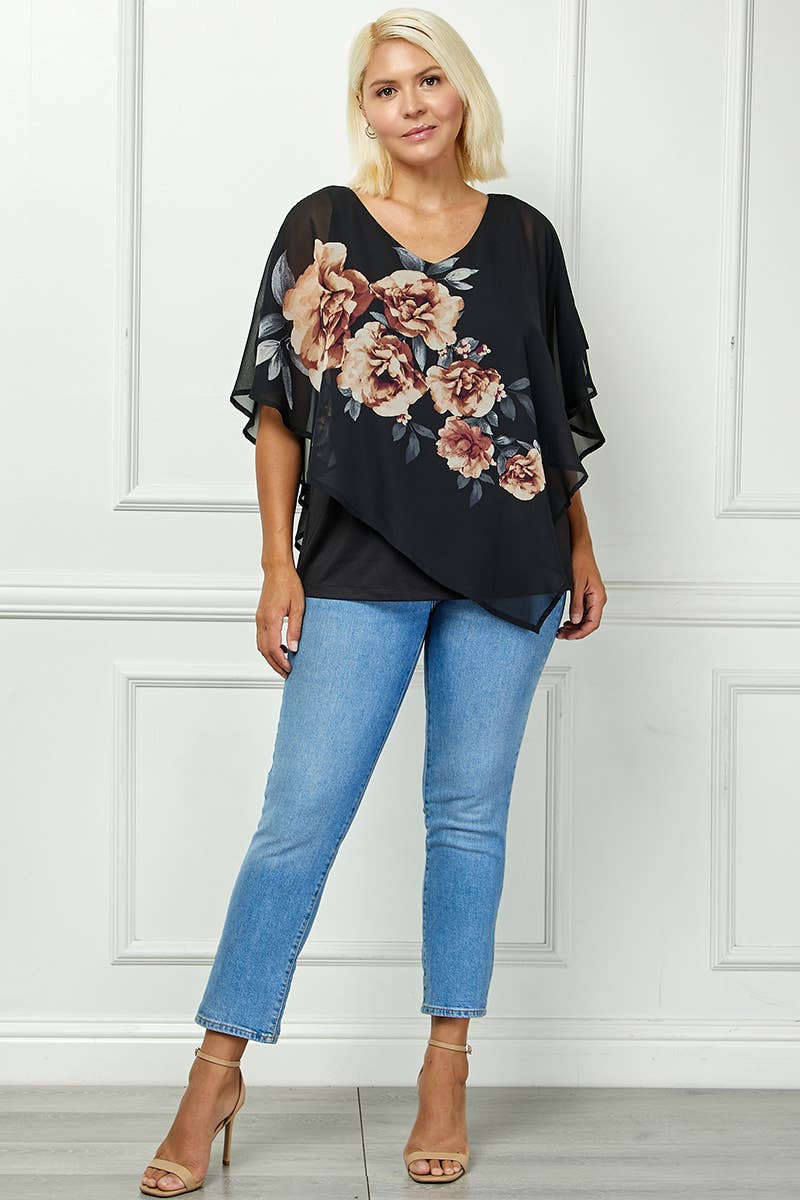 A woman with blonde hair poses indoors in front of a white paneled wall wearing the Plus Blooming Poncho Blouse, light blue jeans, and nude heeled sandals.