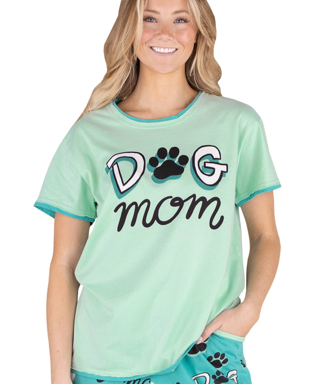 A smiling woman with long blonde hair wears a mint green “Dog Mom” pajama top from the Dog Mom, Cat Nap & More Pajama Tops collection, featuring a paw print in place of the "o." She stands against a white background with one hand in her pocket.
