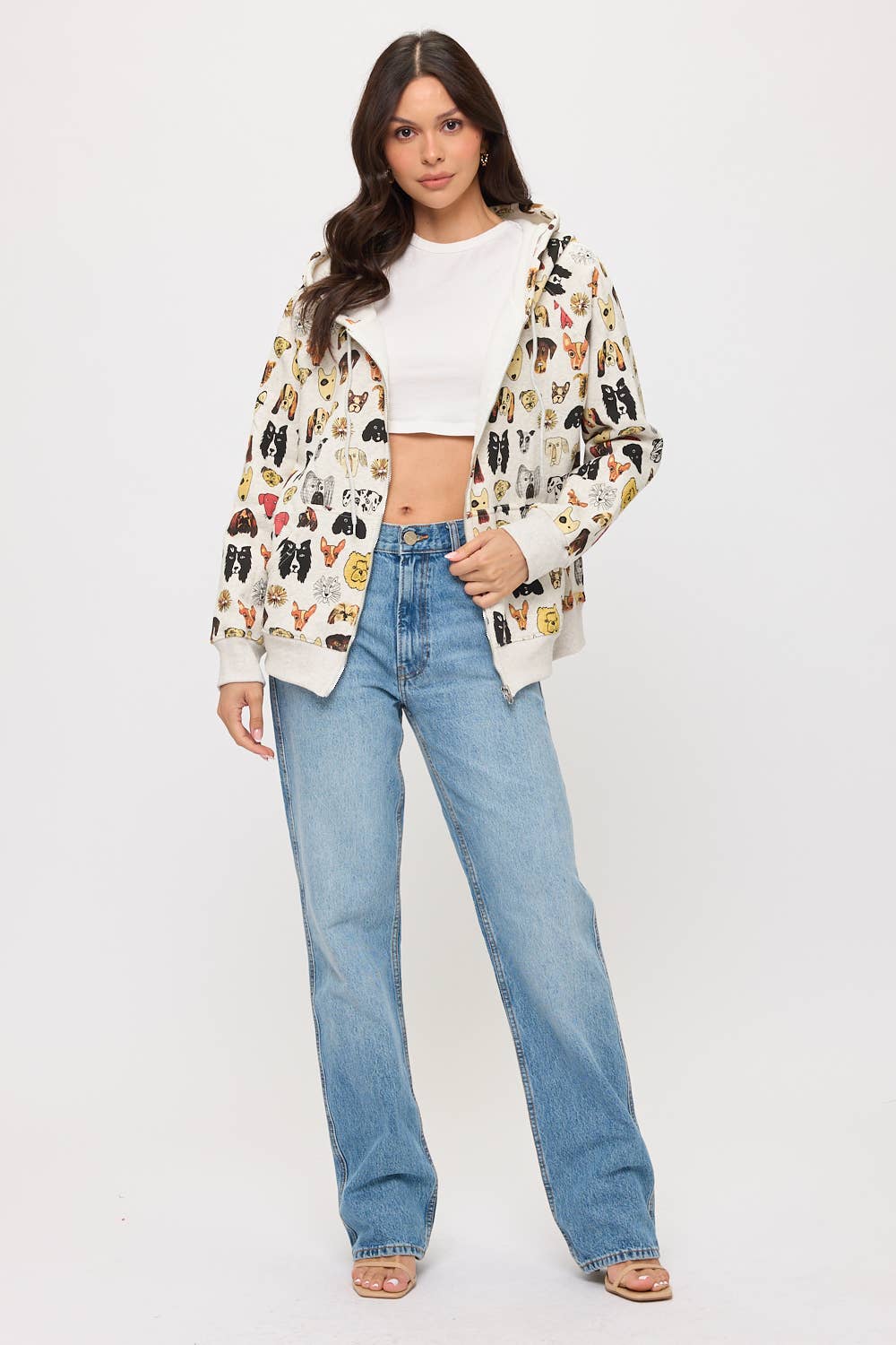 A woman stands against a white background wearing a white crop top, light blue jeans, and the COLORFUL DOG FACES PRINT ZIP UP HOODIE. She has long brown hair, open-toed sandals, and her look is perfect for fans of dog lover apparel.