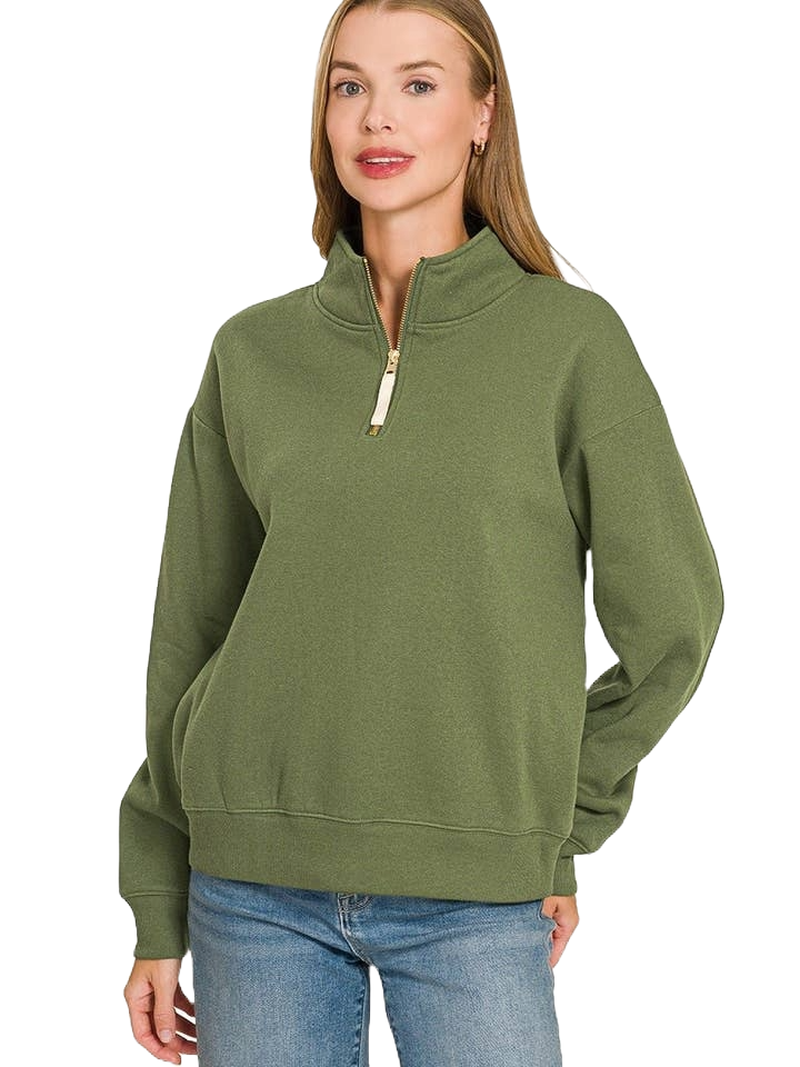 A woman stands facing forward against a white background, smiling with one hand in her pocket, wearing the Fleece Half Zip Sweatshirt in green with a relaxed fit and blue jeans.