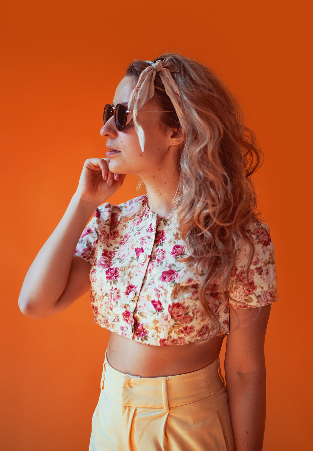 A fair-skinned woman with blonde wavy hair, wearing a floral crop top, yellow pants, and sunglasses, poses against an orange background.
