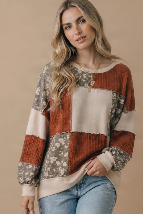 A woman wearing the Plus Color Block Top with floral patchwork in earth tones and blue jeans stands against a beige background, looking at the camera with a relaxed expression and one hand in her pocket.