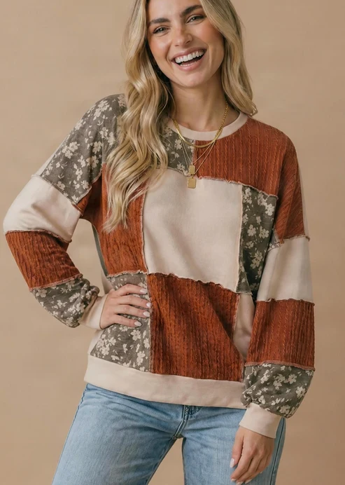 A smiling woman with long blonde hair wears the Plus Color Block Top, which features floral patchwork in brown, beige, and olive tones with color block details. She pairs it with light blue jeans and stands against a plain beige background.