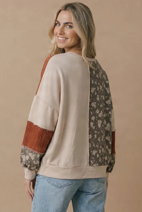 A woman with long blonde hair smiles as she poses in the Color Block Knit Top, a relaxed fit women’s top showcasing both floral knit and solid patterns in a color-blocked design. Paired with light blue jeans, she turns slightly to reveal the back of the top against a beige background.