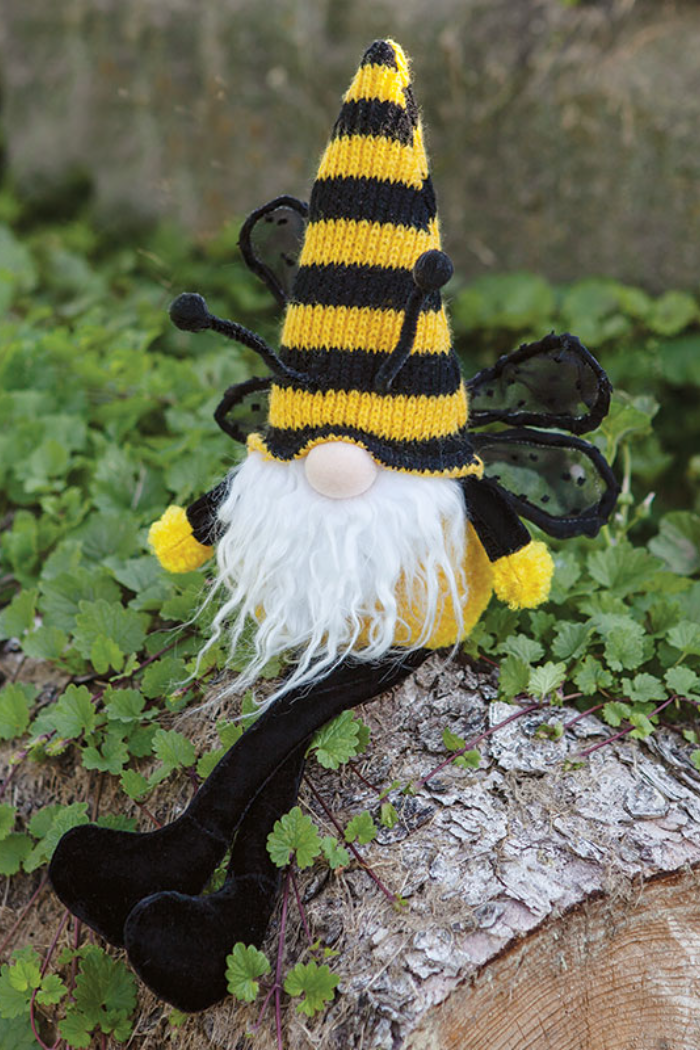 The image shows a "Buzzing Gnome Bee with Dangle Legs." This plush toy gnome is designed to resemble a bee, featuring a black and yellow striped conical hat, small black antennae, and sheer black wings. It has a round, skin-toned nose, a long, white faux fur beard, and black dangling legs with black feet. The gnome's body is yellow, and it has small yellow pom-poms at the ends of its arms. The gnome is posed sitting on a piece of wood, with a background of green foliage.