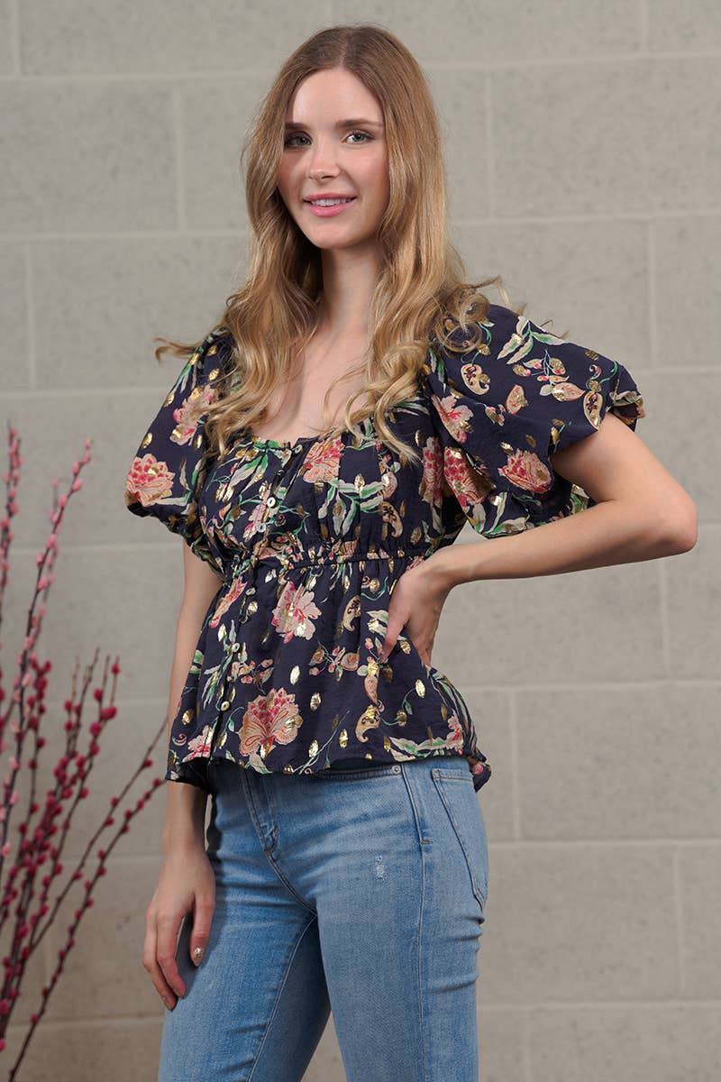 Woman with long blonde hair, wearing a navy-blue floral print blouse. The blouse has short, puffed sleeves, a square neckline, and a cinched waist. The floral pattern features pink, orange, and gold accents.