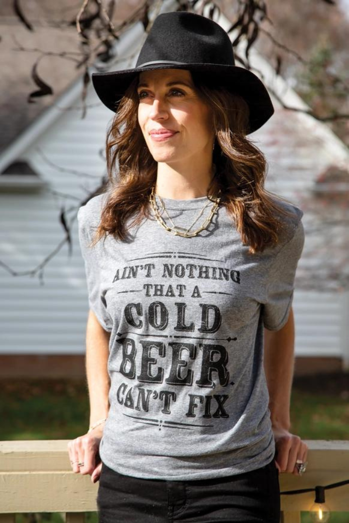 Woman wearing a gray t-shirt with the text "AIN'T NOTHING THAT A COLD BEER CAN'T FIX." She is also wearing a black hat and a gold chain necklace.